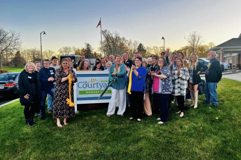 A crowd standing around The Courtyard Memory Care Sign with a cut ribbon